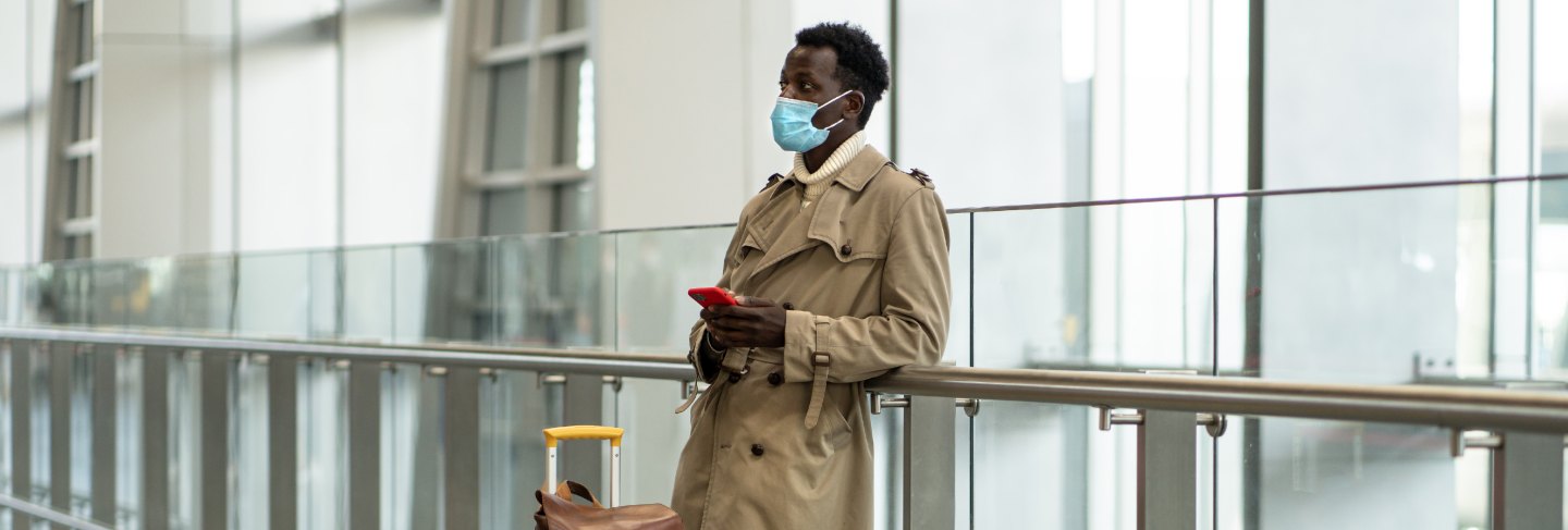 Afro-american traveler man with yellow suitcase stands in airport terminal, wear protective face mask to protect yourself from flu virus, pandemic covid-19, waiting for flight and boarding
